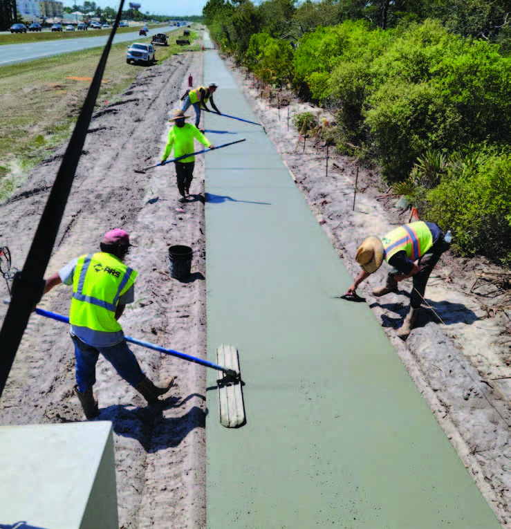 Crew hand-finishing a fresh concrete sidewalk path through a sandy roadside area