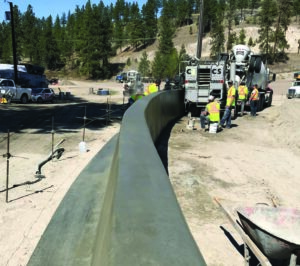 Power Curber 5700-C pouring a curved concrete barrier wall with a mixer truck and forested hills behind