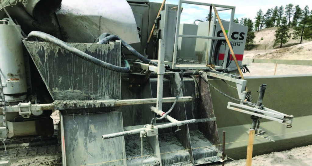 Rear section of a curbing machine coated in wet concrete residue, with hydraulic hoses and a 'CS' concrete services trailer in the background.