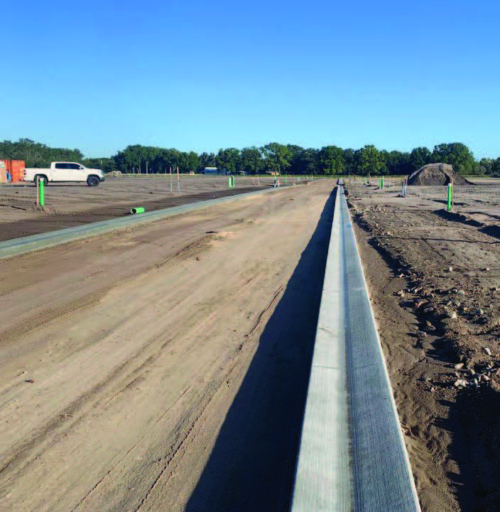 Freshly placed concrete curb running along a dirt roadway under construction, with a pickup truck and grading stakes in the distance.