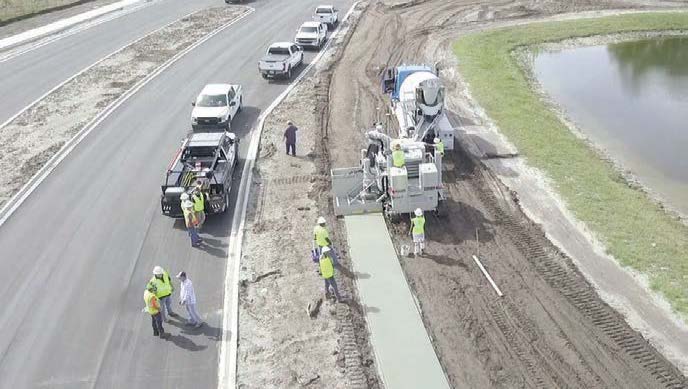 Elevated view of a curbing crew placing concrete along a roadway near a pond, with support vehicles and workers nearby.