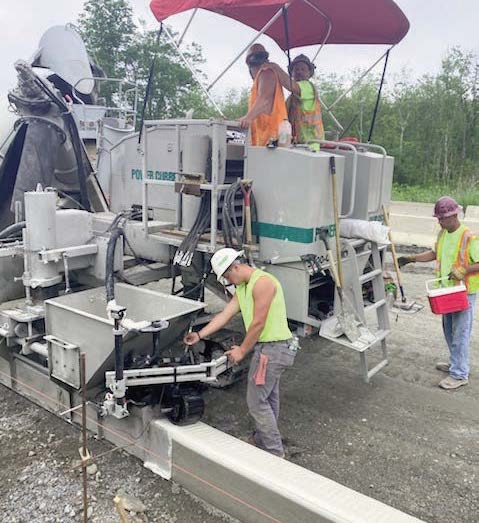 Power Curber machine operating under a red canopy with two workers on the platform and a third worker in a high-visibility vest nearby.