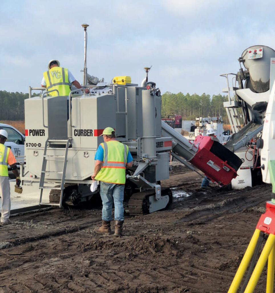 Power Curber 5700-D placing concrete curb as a ready-mix truck pours material, with WG-branded workers on site.