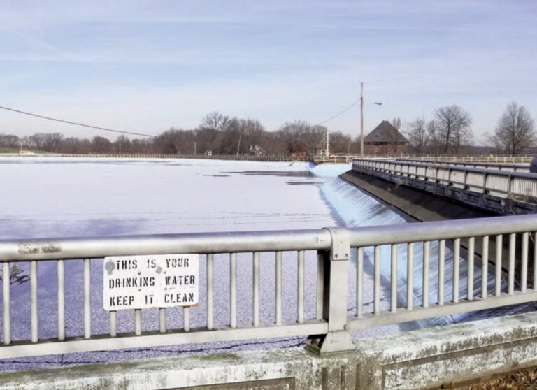 Bridge overlook of a frozen reservoir with a 'This is your drinking water, keep it clean' sign on the railing.