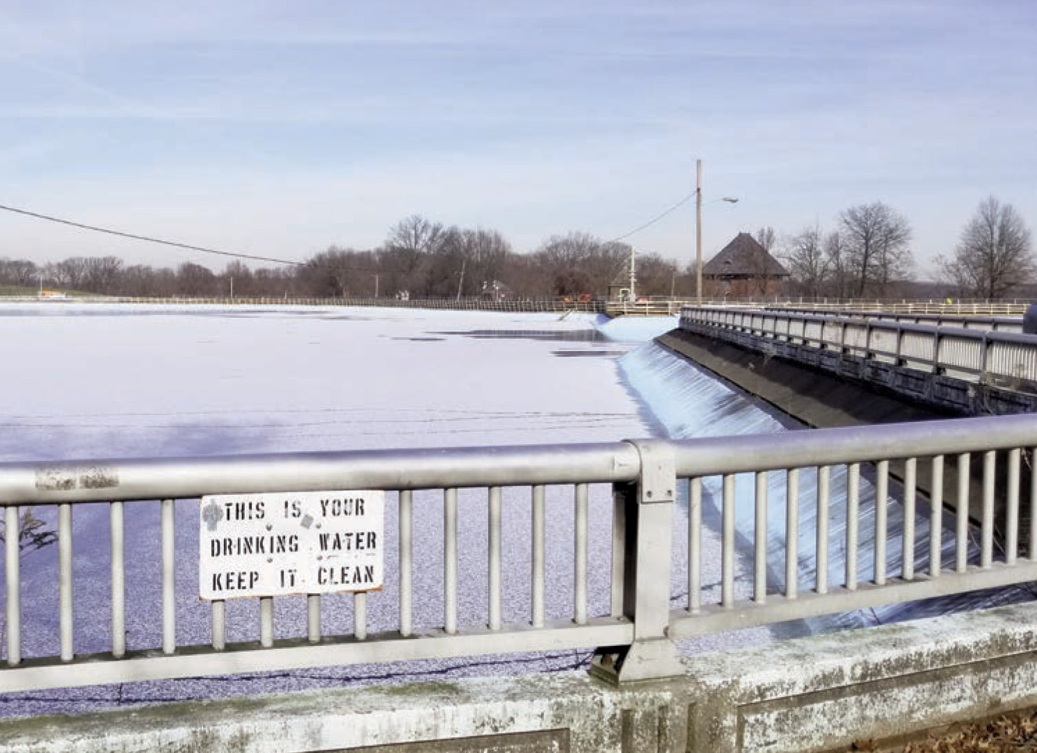 Bridge overlook of a frozen reservoir with a 'This is your drinking water, keep it clean' sign on the railing.