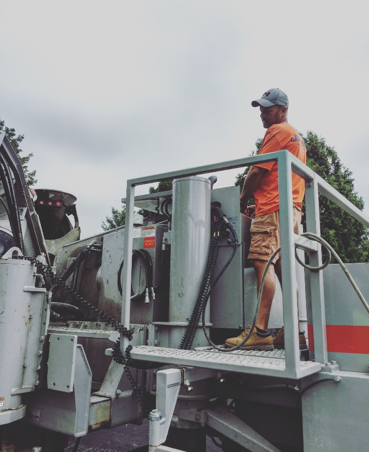 Worker in an orange shirt standing on the hydraulic platform of a curbing machine, adjusting equipment.