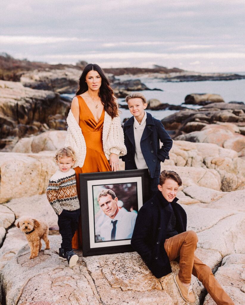 Family portrait on a rocky coastal shoreline with a woman, three children, a dog, and a framed photograph held among them.