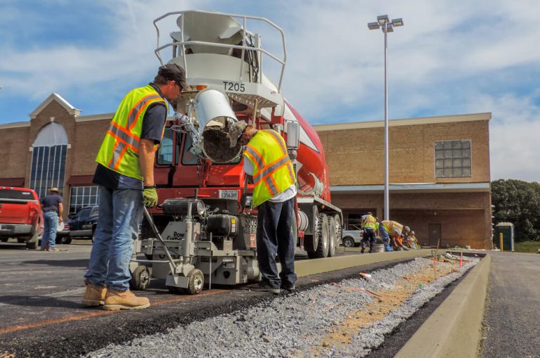 Workers guiding a concrete curb machine as a ready-mix truck pours material in a retail parking lot.