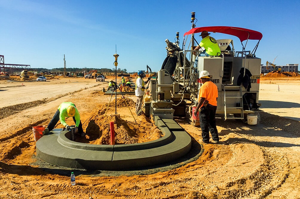 Power Curber 5700-C machine forming a curved concrete curb on a construction site, with a worker in a yellow vest nearby.