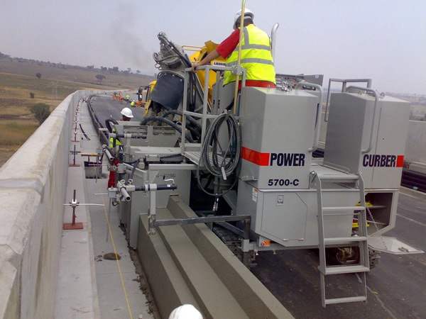 Power Curber 5700-C machine placing a barrier wall on an elevated highway overpass, with a worker in high-visibility gear.