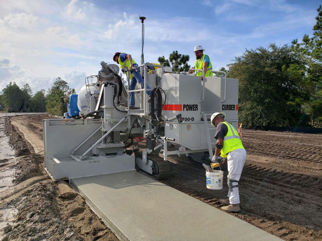 Power Curber 5700-D machine placing a concrete slab as two workers in high-visibility vests operate controls and a worker in white finishes the surface.