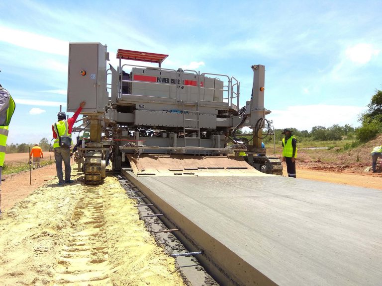 Power Curber machine paving a concrete road on a red-dirt site in Africa, with workers in high-visibility vests guiding the pour.