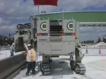 Worker in an orange vest standing beside a Power Curber 5700-C machine with CB branding, with a green storage tank in the background.