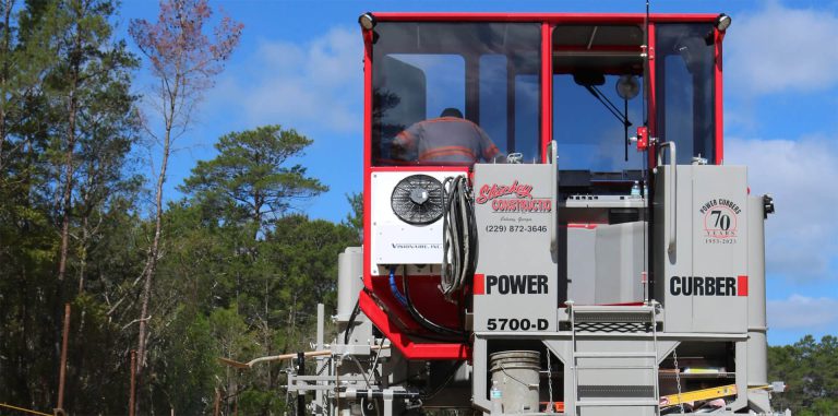 Enclosed, climate-controlled cab of a Power Curber 5700-D machine bearing Stuckey Construction and Power Curbers 70th-anniversary decals, with an operator inside.