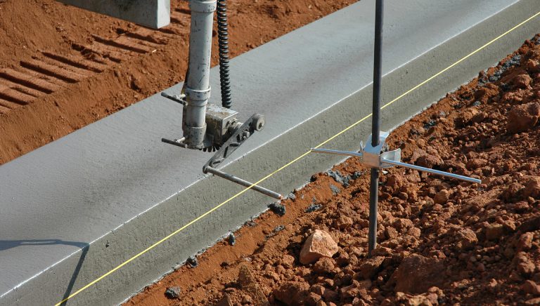 Close-up of a curbing machine's slipform mold and guidance sensor tracking a stringline beside a freshly placed concrete curb on a red-dirt site.