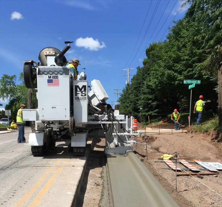 Miller Formless M-1000 curb and gutter machine with an American flag decal placing curb along a roadway as workers look on near a street sign.