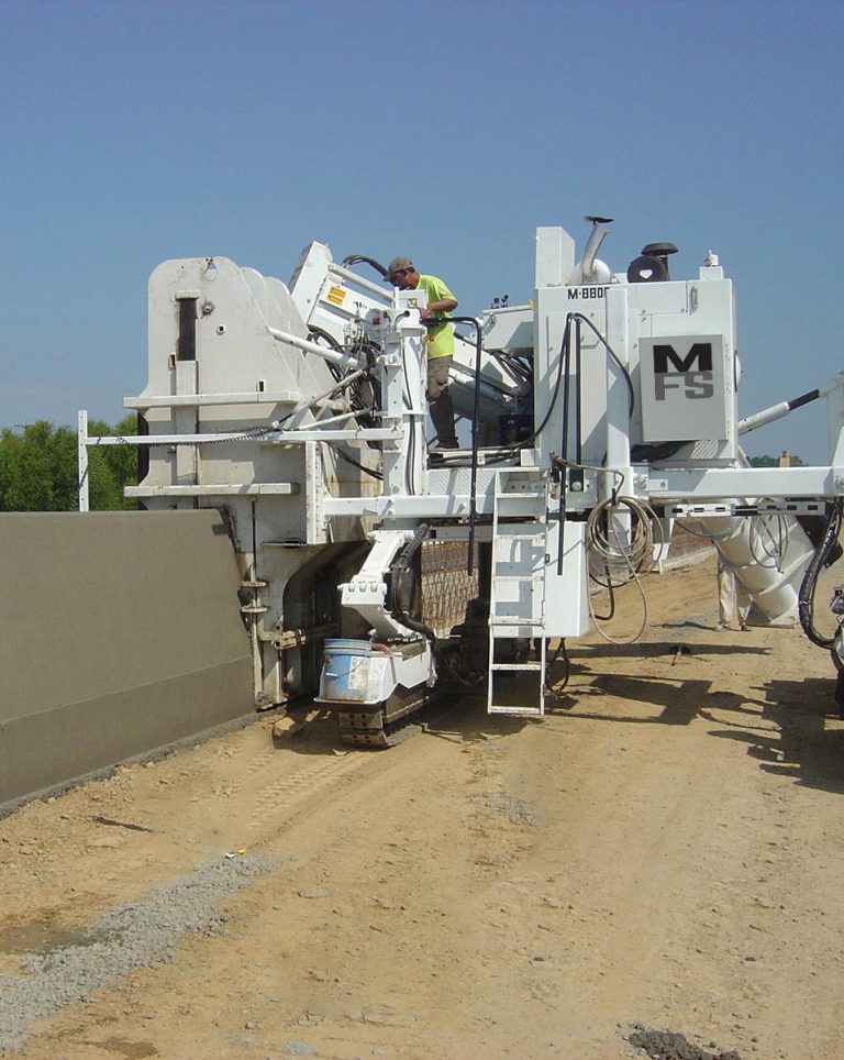 Miller Formless M-8800 machine forming a concrete barrier wall, with a worker in a high-visibility shirt monitoring the equipment.