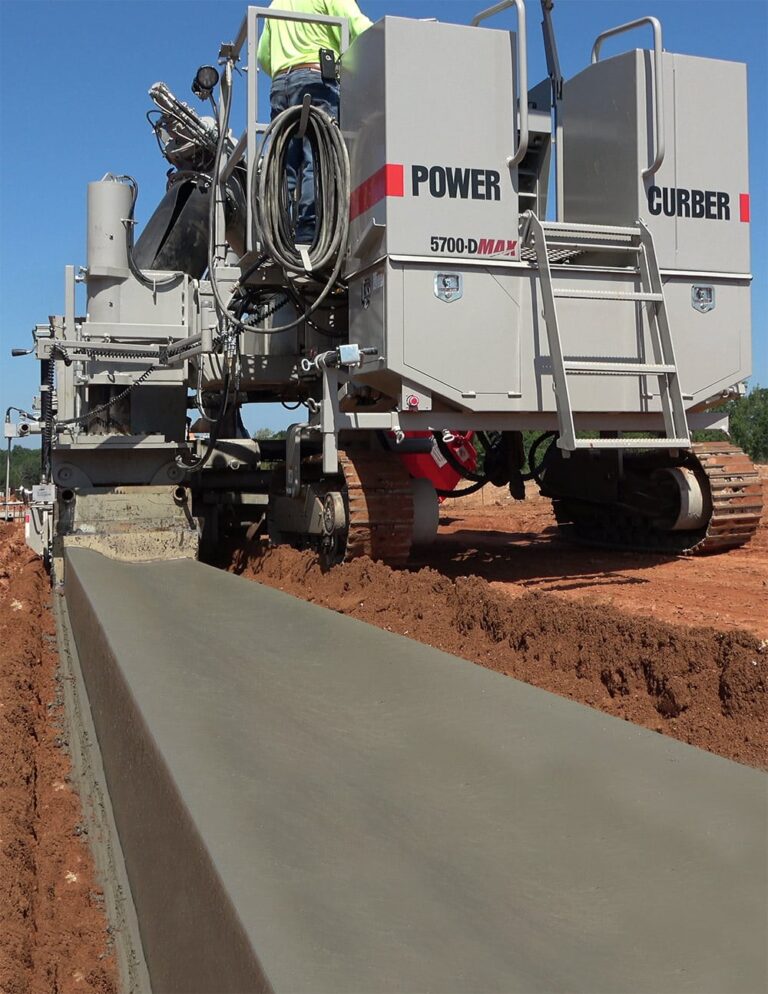 Low-angle view of a Power Curber 5700-D MAX machine placing concrete curb on a red-dirt site, with a worker in a high-visibility vest on the platform.