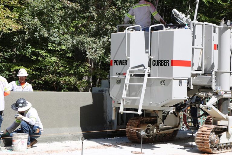 Power Curber 5700-D MAX machine forming a concrete parapet wall as a worker finishes the surface by hand.