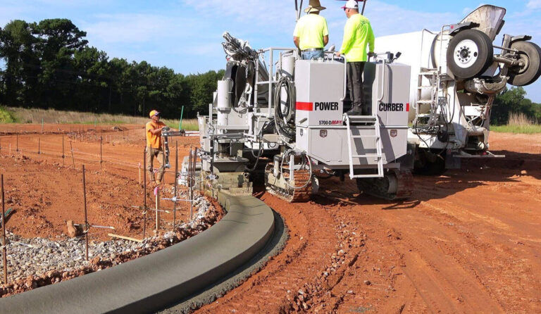Power Curber 5700-D MAX machine placing a curved concrete curb, with two workers on the platform and a ready-mix truck feeding material.