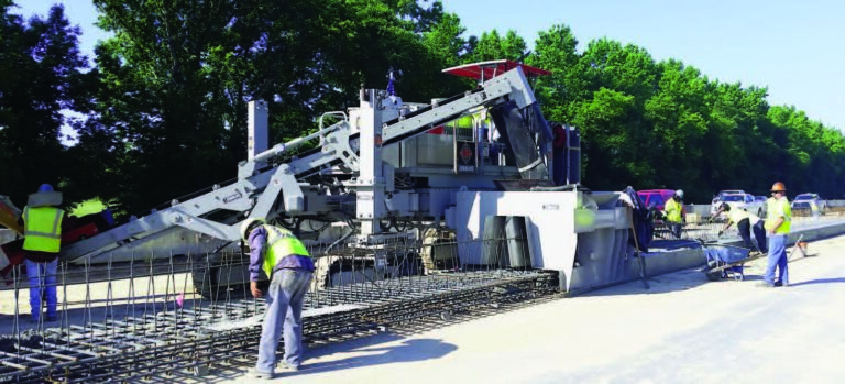 Slipform machine with COMMAND branding forming a reinforced concrete barrier wall on a highway median, with workers placing rebar and material.