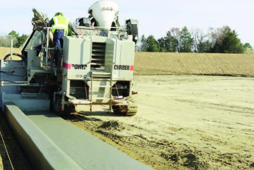 Power Curber machine placing concrete curb as a ready-mix truck feeds material, with a worker in a high-visibility vest and an open field in the background.