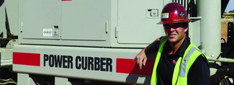 Worker in a red hard hat and safety vest standing beside a Power Curber machine's equipment housing.