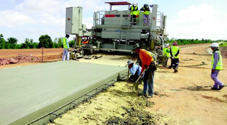 Power Curber 7700 machine paving a wide concrete roadway with several workers guiding the pour and finishing the surface.
