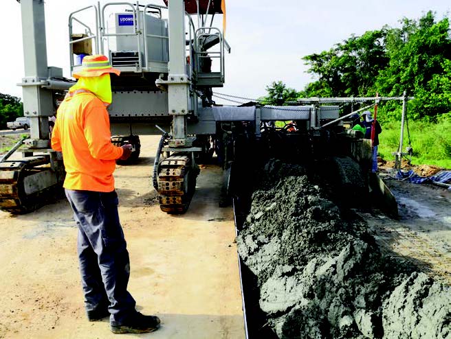 Worker in high-visibility gear operating a remote control beside a slipform machine placing concrete into a barrier wall form.