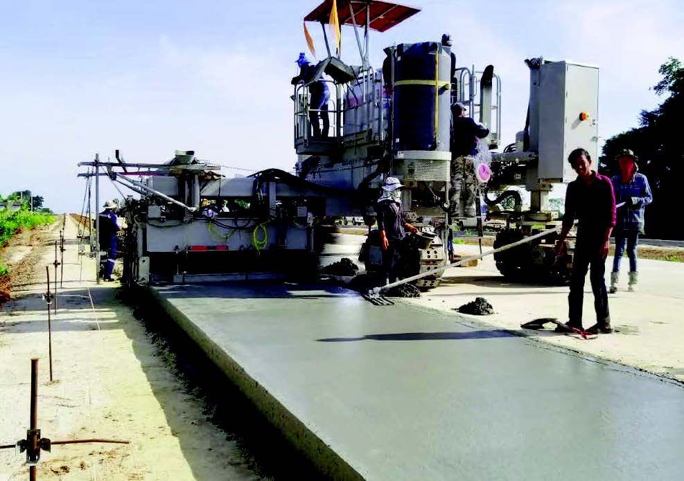 Slipform curbing machine paving a concrete road under a red canopy, with workers using a straightedge tool to finish the surface.