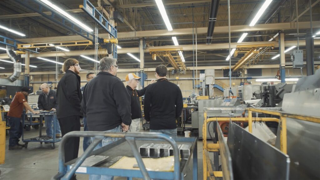 Group of workers touring a manufacturing shop floor with overhead cranes and metal fabrication equipment during a training session.
