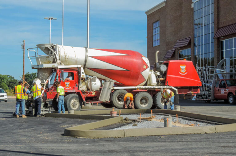 Ready-mix concrete truck pouring material as workers in high-visibility vests finish a curbed planting bed outside a retail building.