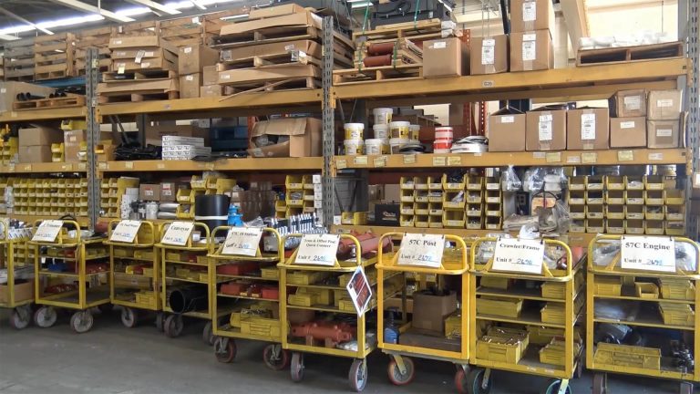 Warehouse shelving stocked with labeled parts carts for Power Curber machine components, including trimmer, conveyor, post, and engine assemblies.