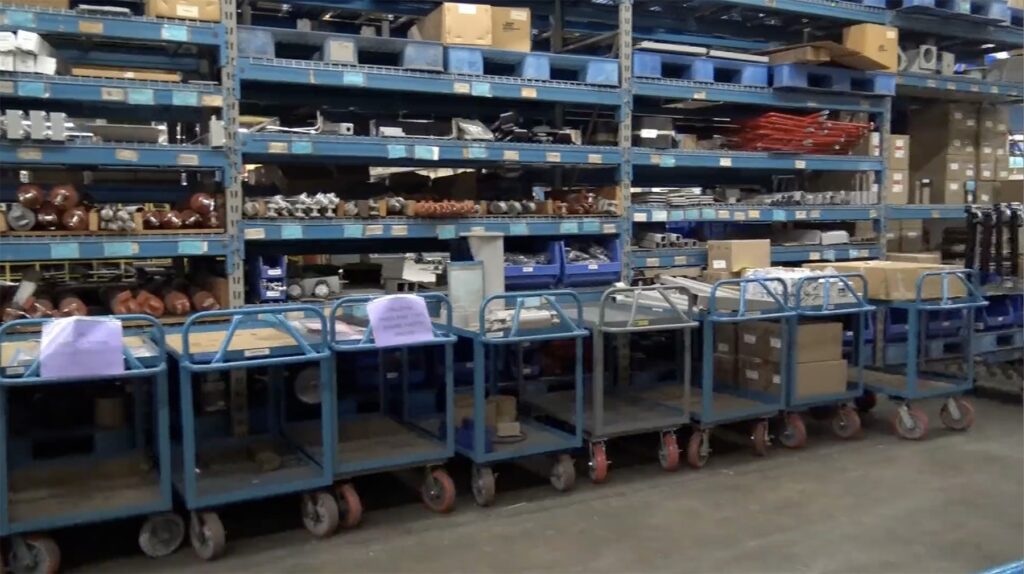 Warehouse shelving and rolling carts holding spare machine parts and hardware bins.