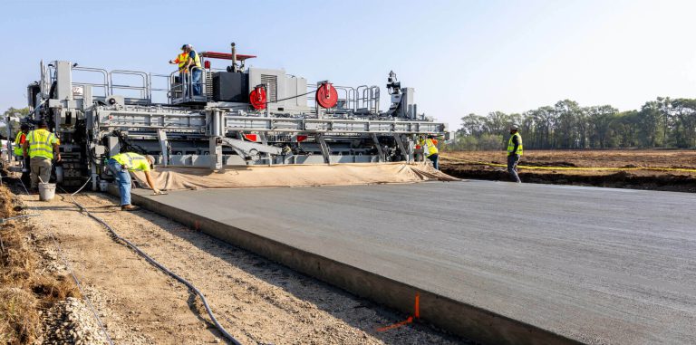 Power Pavers slipform machine placing a wide concrete slab as workers guide the pour and finish the surface on an open site.