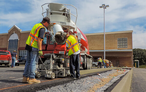 Power Curber S809 machine placing curb as a ready-mix truck feeds concrete, with two workers guiding the pour near a retail store.