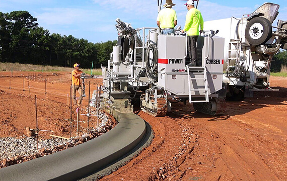 Power Curber 5700-D MAX machine placing a curved concrete curb on a red-dirt site, with two workers on the platform and one guiding the stringline.