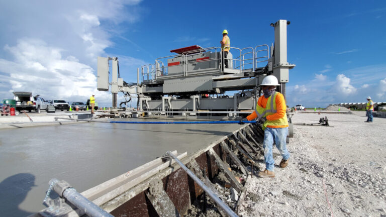 Power Curber 7700 machine placing a wide concrete slab on an airport apron, with a worker finishing the edge and support vehicles nearby.