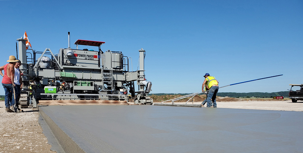 Power Paver SF-2404 placing a wide concrete slab as workers finish the surface with a straightedge on a sunny airport site.