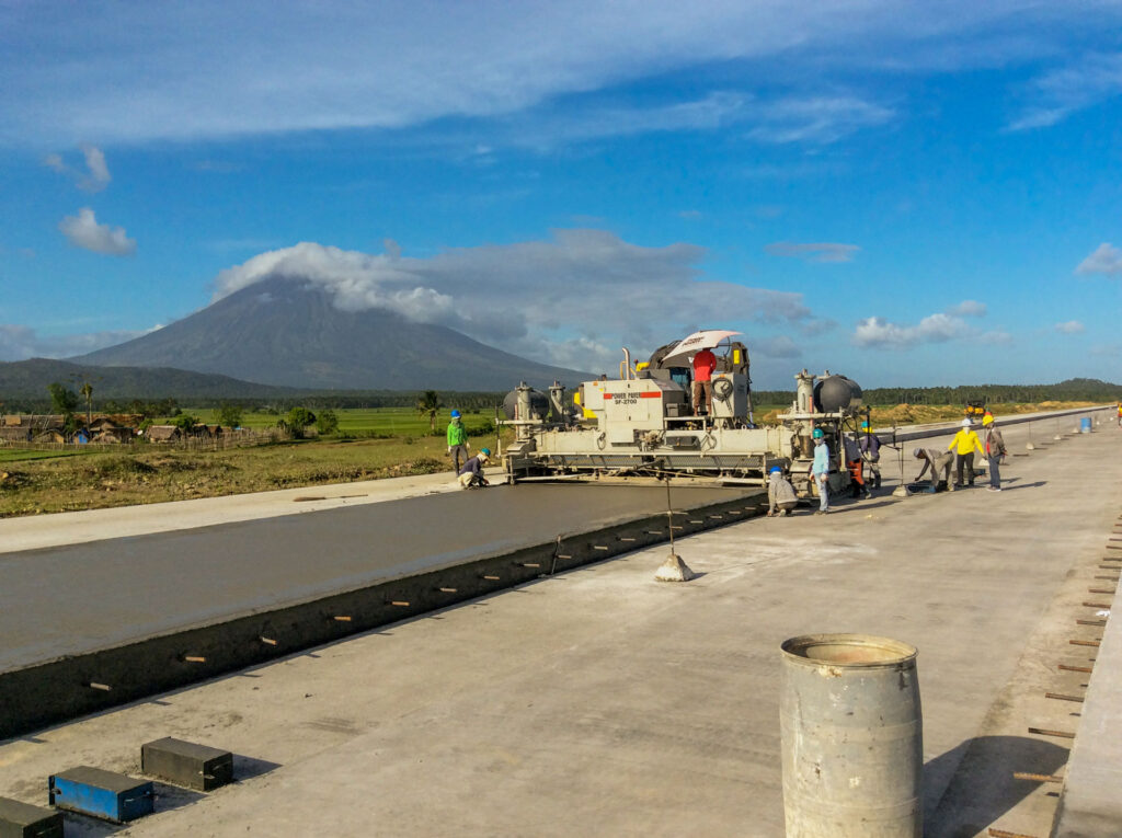 Power Paver SF-2700 placing a concrete road slab with a volcano in the background and workers finishing the surface.