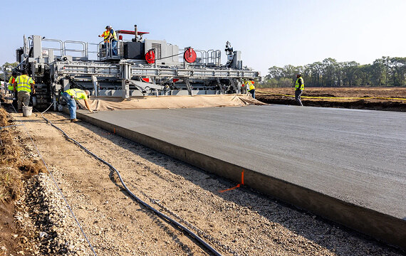 Power Paver SF-3404 slipform paver placing a wide concrete slab as workers finish the surface on a dirt job site.