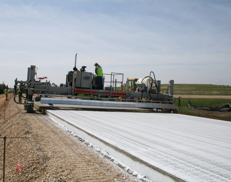 Power Paver TC-2700 texture and curing machine finishing a wide concrete road slab, with a worker on the platform.