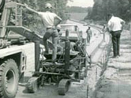 Black-and-white archival photo of a Power Curbers machine placing curb along a roadway lined with trees.