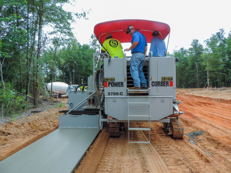 Power Curber 5700-C machine with a red canopy placing a wide concrete slab, with a mixer truck and three workers on the platform.