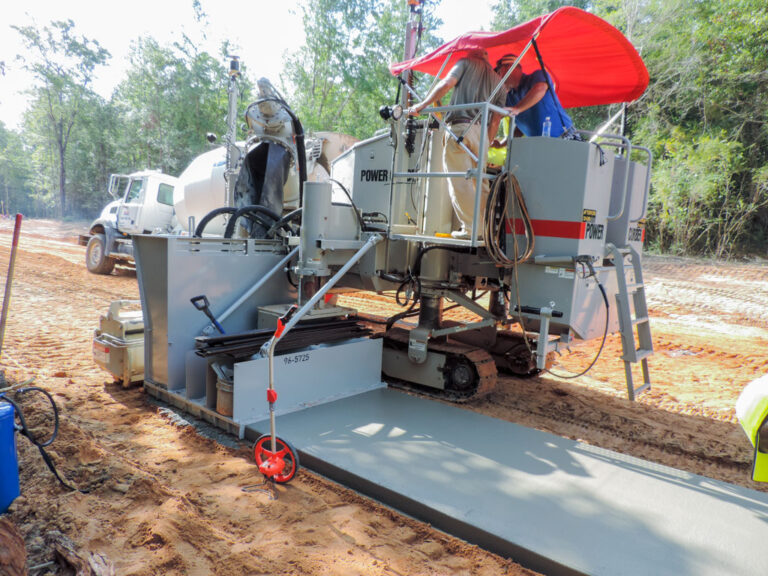 Power Curber machine with a red canopy placing a concrete slab, with a mixer truck feeding material and two workers on the platform.