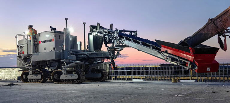 Power Curber 5704 machine with an auger attachment at dusk, with a worker on the platform and an excavator feeding material.
