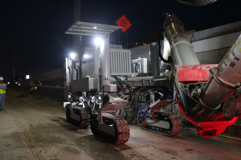 Power Curber machine with an auger attachment placing a concrete barrier wall at night under work lights, with a worker in a high-visibility vest nearby.