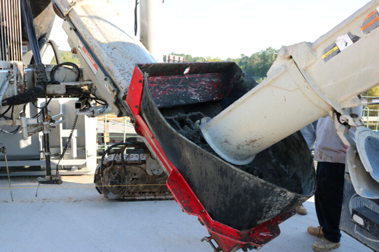 Close-up of a Power Curber auger hopper feeding wet concrete into a placer boot as a worker looks on.