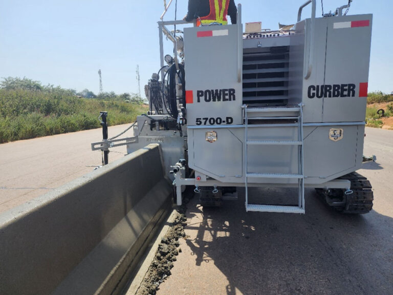 Rear view of a Power Curber 5700-D machine placing a concrete barrier wall along a roadway, with a worker on the platform.