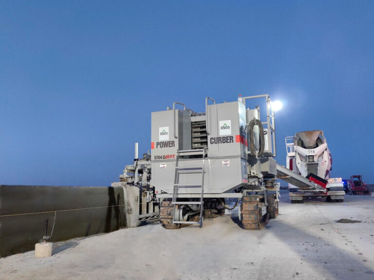 Power Curber 5704-D MAX machine branded RM09 placing a barrier wall at dusk, with a mixer truck feeding material under work lights.